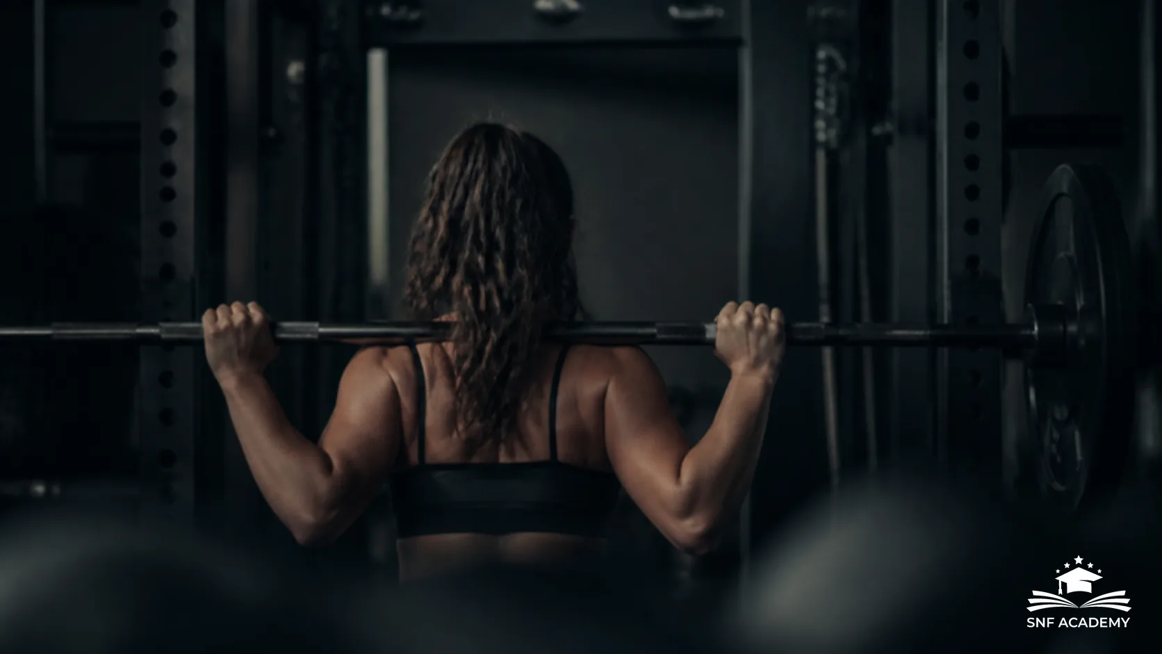 Frau von hinten greift eine Langhantel am Squat-Rack im Krafttraining